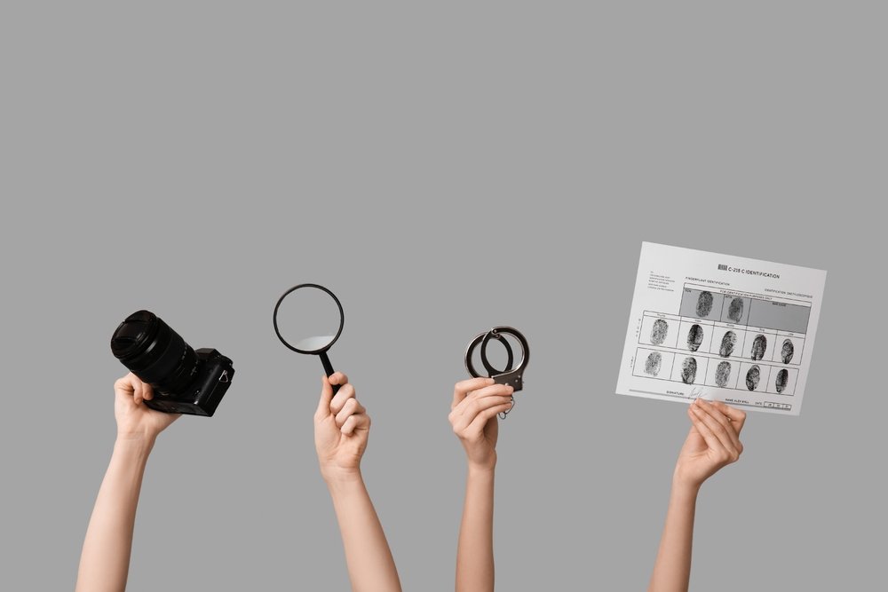 Braços segurando elementos que remetem ao trabalho de um Detetive Particular, sendo uma câmera, uma lupa, um par de algemas e uma folha de digitais impressas.